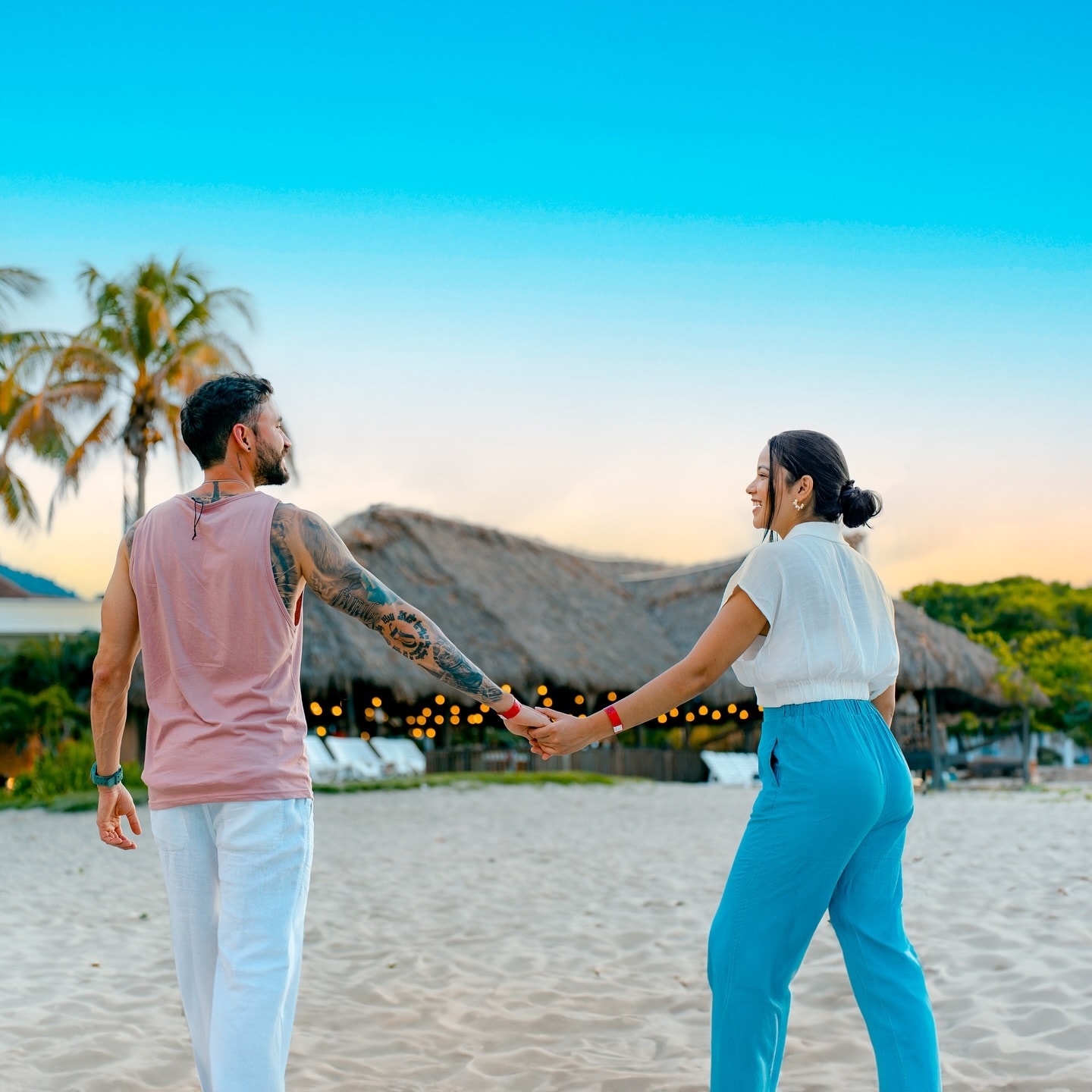 Pareja caminando de la mano por la playa al atardecer, frente a una cabaña de palma